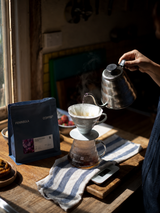 Person pouring coffee from a kettle into a filter on a wooden table with a window in the background.