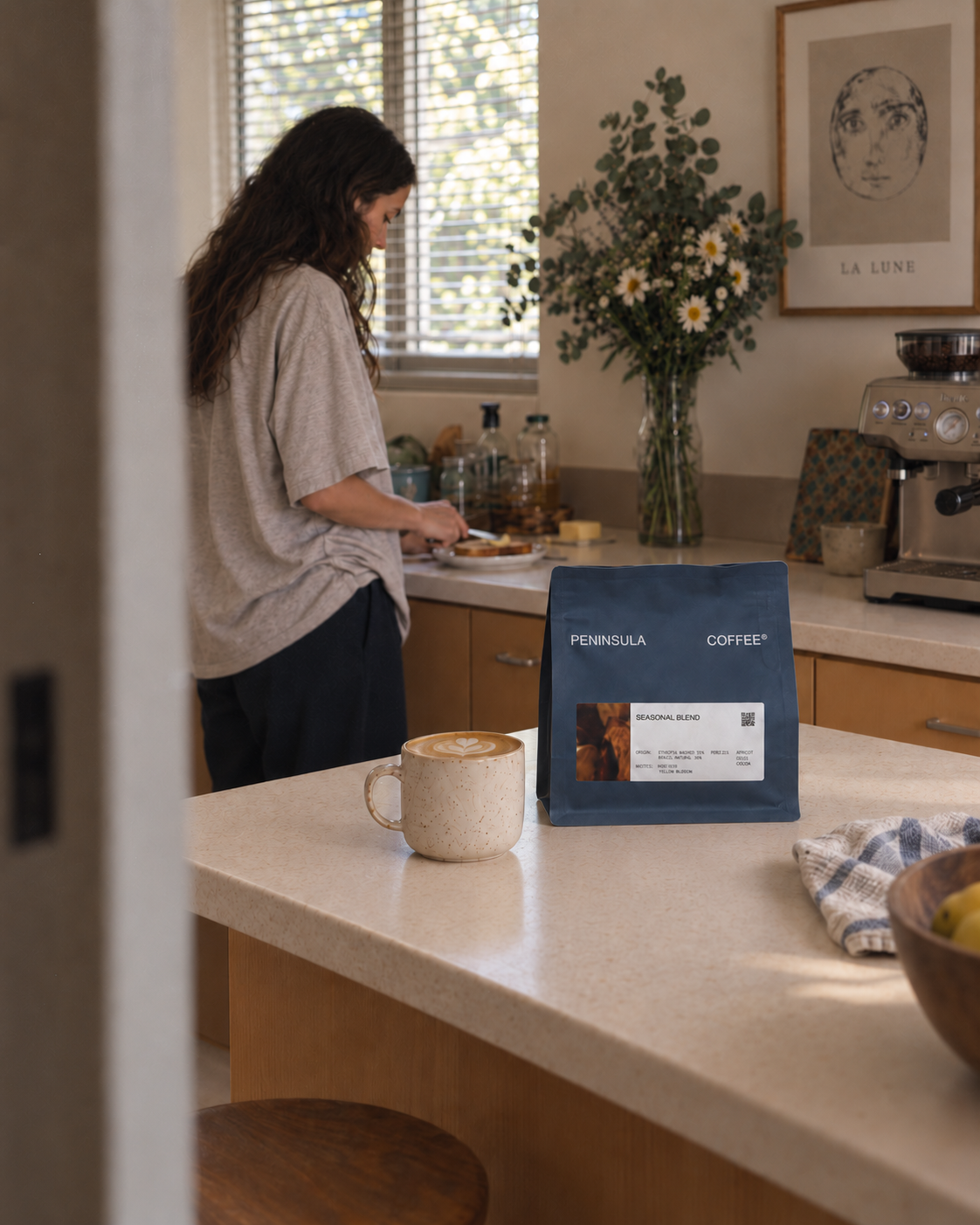 Peninsula Coffee Seasonal Blend beside a flat white on a kitchen bench, with a woman making breakfast in the background.