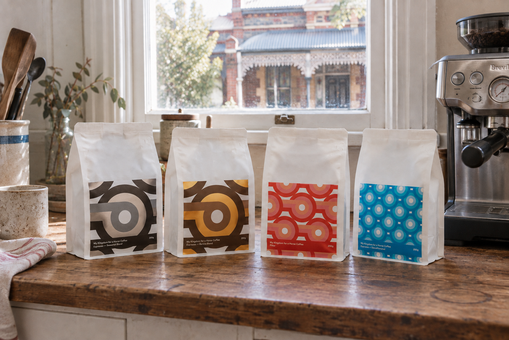 Four My Kingdom for a Horse coffee bags lined up on a timber kitchen bench beside a Breville espresso machine.