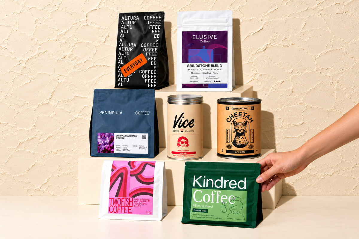 Seven Adelaide roasters' coffee packs centred on cream steps against textured plaster, with a hand placing the Kindred bag.